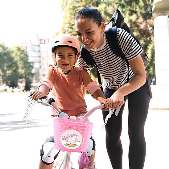 fille a velo equiper de proteges genoux et coude blancs, casque velo rose et velo panier rose personnaliser licorne assister par mere portant sac a dos
